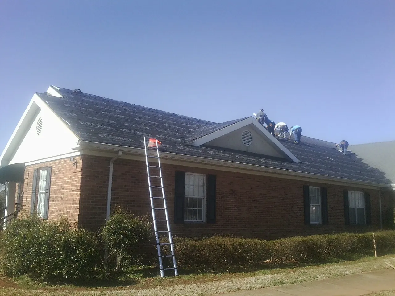 Crew tearing shingles off a brick ranch roof with a ladder set — Metal Roof Repair work by our crew in Mount Blanchard