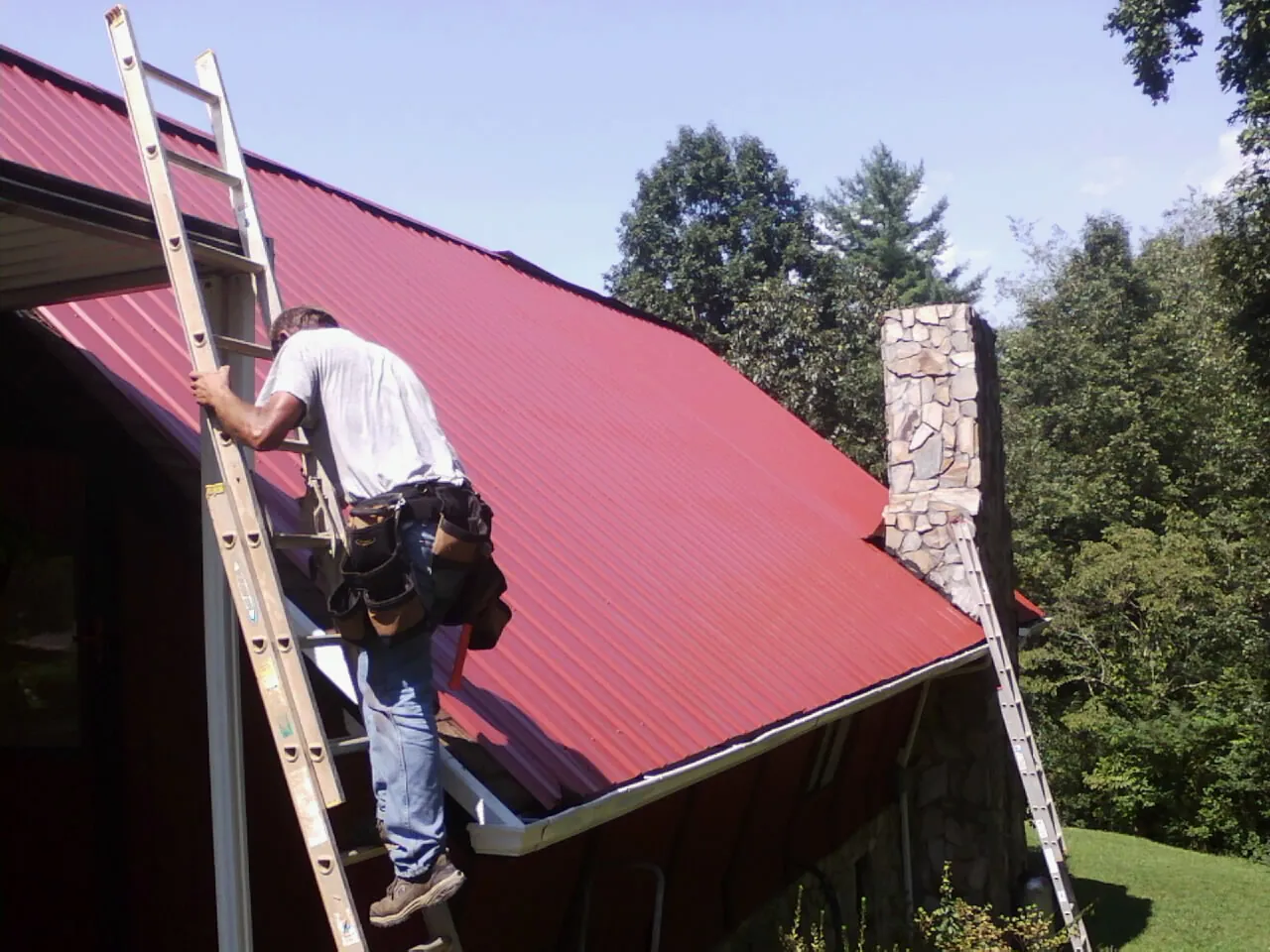Roofer on a ladder beside a red metal roof and stone chimney — Soffit & Fascia Repair by Roofing Brothers in Mount Blanchard